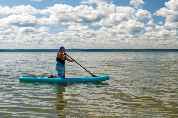 Una mujer en traje de baño cerrado, un pareo y un tocado de rodillas en una tabla de SUP con un remo flota en el agua