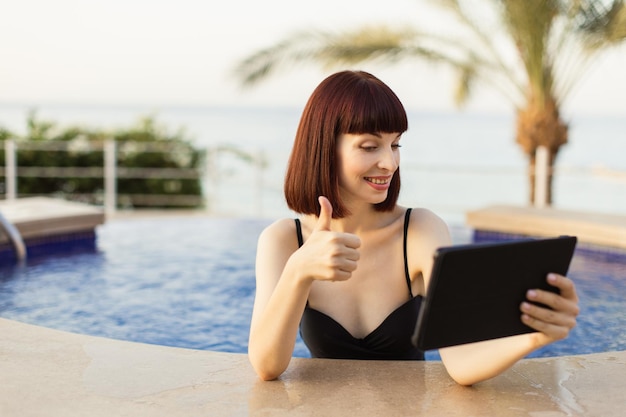 Mujer con traje de baño negro usando tablet pc y mostrando el pulgar hacia arriba mientras nada en la piscina