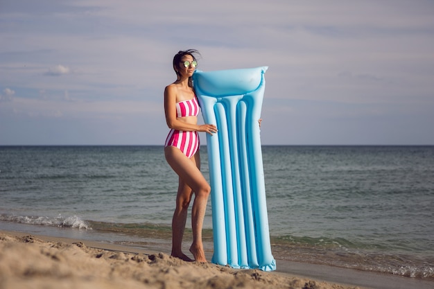 Mujer en traje de baño de rayas rosa se encuentra en una playa de arena con colchón inflable azul