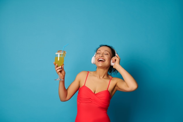 Mujer en traje de baño rojo y auriculares sosteniendo un cóctel en la mano sobre fondo azul.