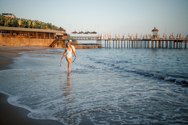 Una mujer en traje de baño y un sombrero con gafas camina por la playa al atardecer. El concepto de recreación en el mar. Enfoque selectivo