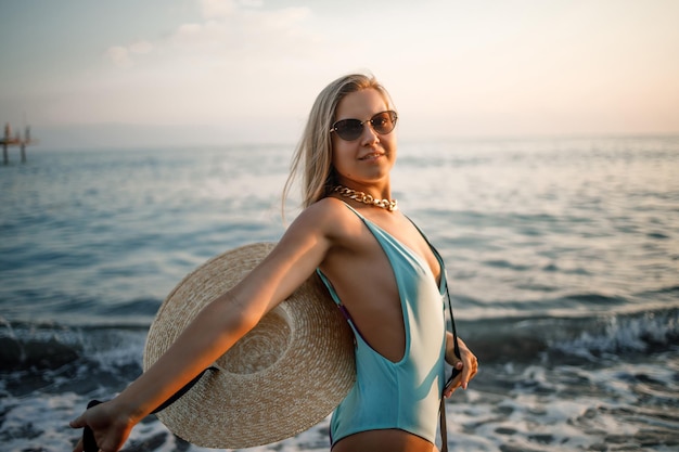 Una mujer en traje de baño y un sombrero con gafas camina por la playa al atardecer. El concepto de recreación en el mar. Enfoque selectivo