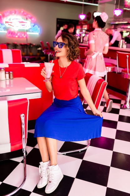 Mujer en traje colorido en café retro de los años 50 sentado en la mesa bebiendo un cóctel de batido de leche con falda azul y camisa roja con gafas de sol divirtiéndose en un estado de ánimo alegre