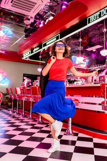 Mujer en traje colorido en retro vintage 50's cafe bailando con chaqueta, falda azul y camisa roja, gafas de sol divirtiéndose en un estado de ánimo alegre