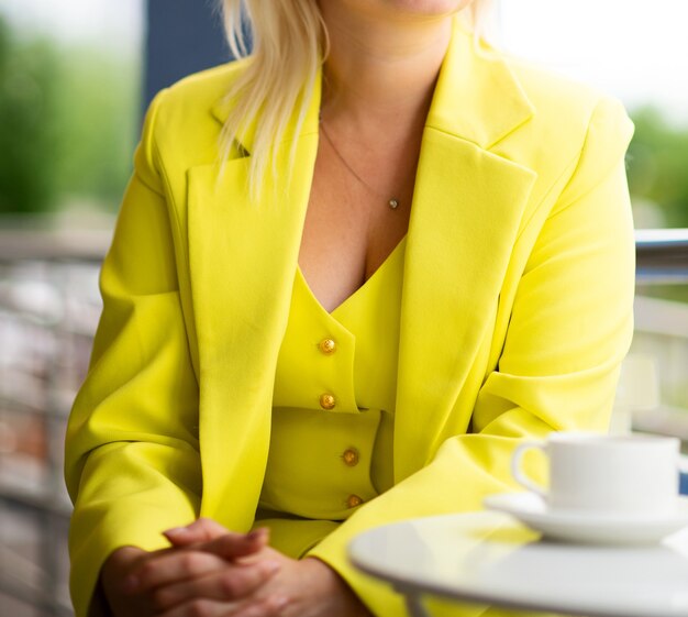 Mujer en un traje de negocios amarillo brillante en una mesa en un café de verano, irreconocible