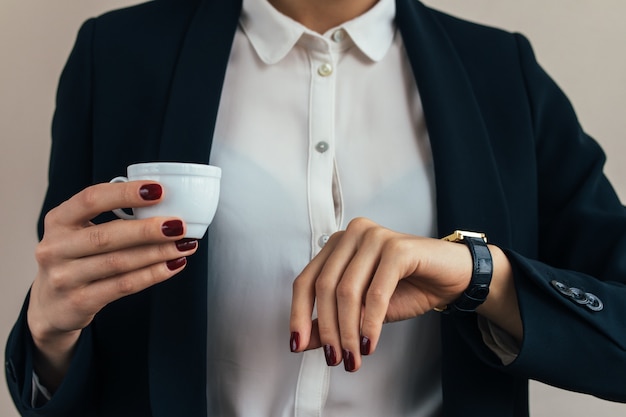 Mujer en un traje de negocios que sostiene una taza de café y comprueba la hora en un reloj de pulsera