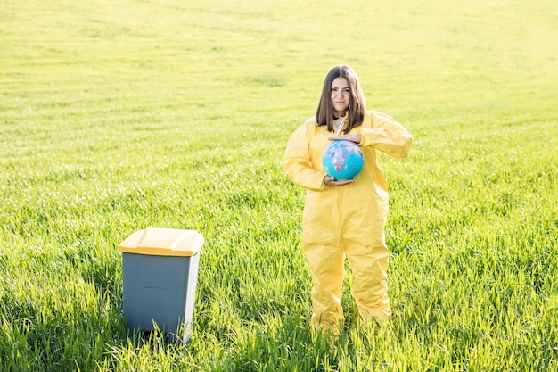 Una mujer con un traje protector amarillo se para en medio de un campo verde y sostiene un globo en sus manos junto a un bote de basura