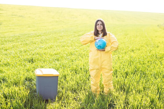 Una mujer con un traje protector amarillo se para en medio de un campo verde y sostiene un globo en sus manos junto a un bote de basura
