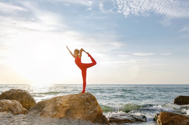 La mujer con un traje rojo practicando yoga en piedra al amanecer cerca del mar