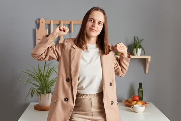 Una mujer tranquila y atractiva con el pelo castaño y una chaqueta beige parada en el interior de la cocina de casa estirando las manos despertándose temprano mirando la cámara