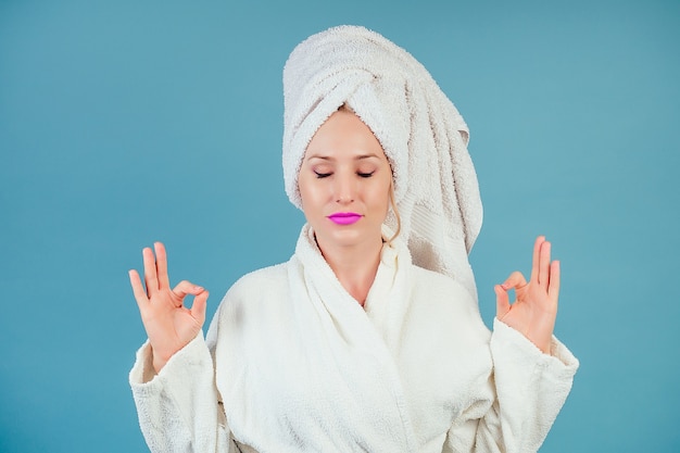 Mujer tranquila atractiva y sonriente en una bata de baño de algodón blanco y un turbante de toalla en la cabeza medita en el estudio sobre un fondo azul copyspace. concepto de spa y relajación.