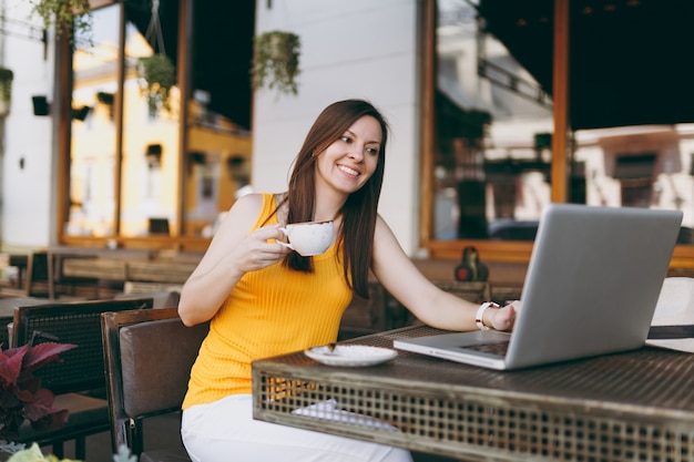 Mujer tranquila en la cafetería de la calle al aire libre sentado en la mesa trabajando en una computadora portátil moderna, beber té, relajarse en el restaurante durante el tiempo libre