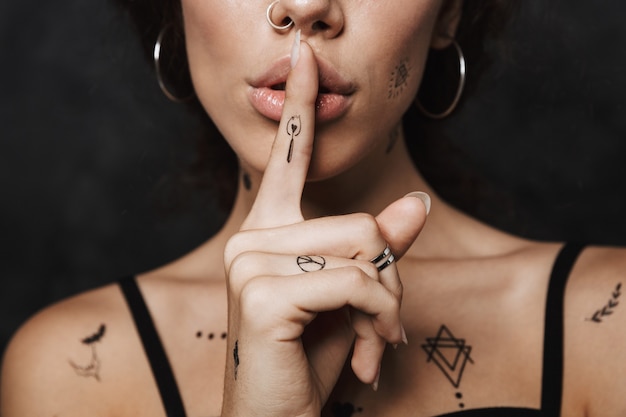Mujer tranquila joven recortada con tatuaje gesticulando signo de silencio con el dedo aislado sobre la pared negra