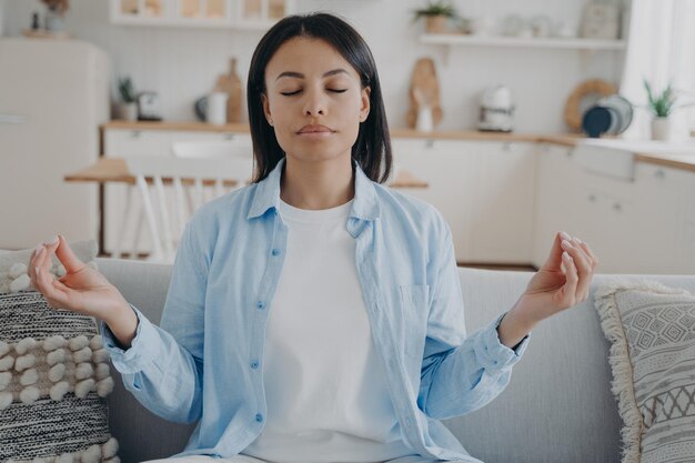 Una mujer tranquila practica yoga meditando con los dedos doblados en un gesto de mudra sentado en el sofá de casa