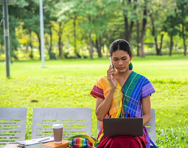 Foto mujer transgénero con un vestido de arcoíris sentada en un banco del parque usa tu teléfono y computadora portátil concepto de estilo de vida lgbtq mes del orgullo lgbtq
