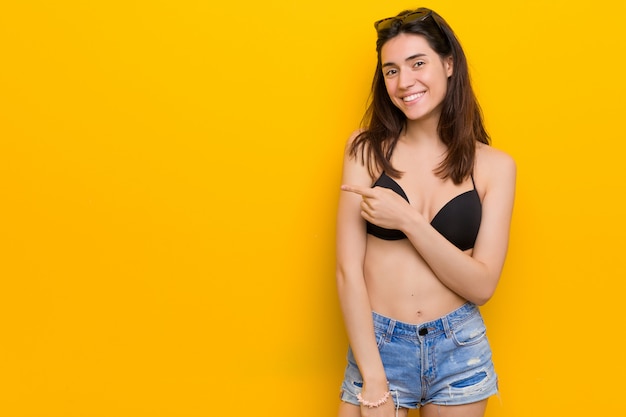 Foto mujer triguena joven que lleva un bikini contra la pared amarilla que sonríe y que señala a un lado, mostrando algo en el espacio en blanco.