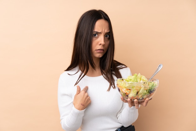 Mujer triguena joven que sostiene una ensalada sobre la pared aislada que se señala