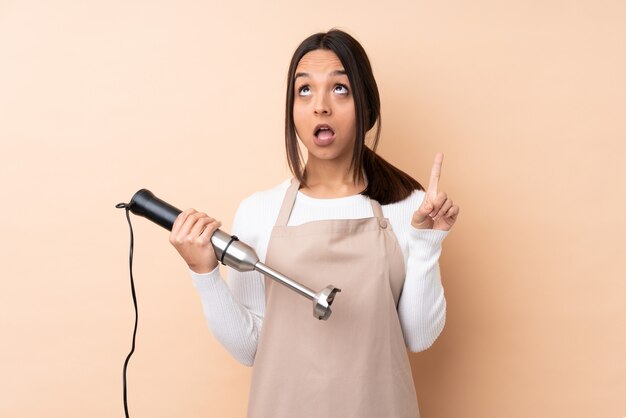 Mujer triguena joven que usa la licuadora de la mano sobre la pared aislada que piensa una idea que destaca el dedo