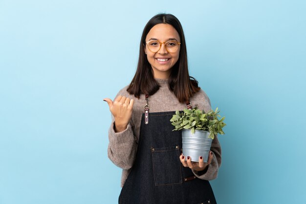 Mujer triguena joven de la raza mixta que sostiene una planta sobre el azul aislado que señala al lado para presentar un producto.