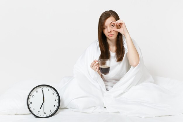 Mujer triste cansada sentada en la cama con una taza de café, reloj redondo, sábana blanca, almohada, envolviendo en una manta en la pared blanca