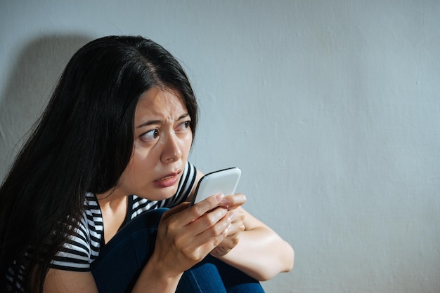 mujer triste e infeliz que encuentra violencia familiar usando un teléfono móvil buscando ayuda en un fondo de pared blanco oscuro que muestra el concepto de mujeres maltratadas maltratadas.