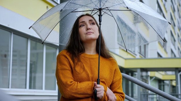 Mujer triste se encuentra bajo un paraguas transparente bajo la lluvia en el fondo de un edificio de apartamentos.