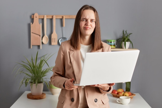 Mujer triste insatisfecha con cabello castaño con chaqueta beige parada en el interior de la cocina trabajando en una laptop mirando la cámara con tristeza en la presentación fallida en línea