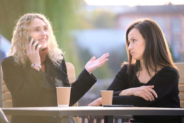 Mujer triste siendo ignorada por su amiga sentada en un café de la calle al aire libre mientras habla alegremente por teléfono móvil y no presta atención. Concepto de problemas de amistad