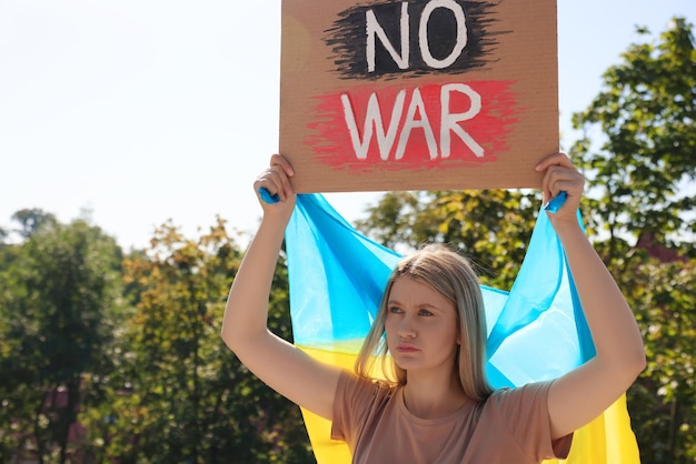 Mujer triste sosteniendo un cartel con las palabras No War y la bandera ucraniana al aire libre