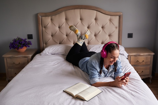 Mujer tumbada en la cama escuchando musica con auriculares junto a un libro