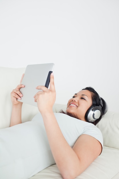 Mujer tumbada en un sofá con auriculares y sosteniendo una tableta táctil