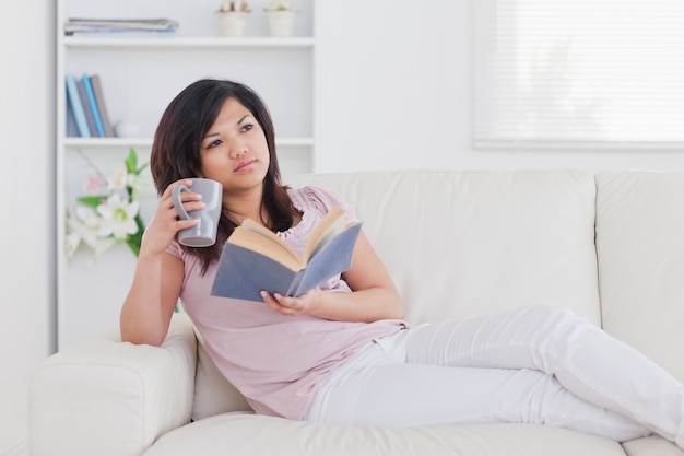 Mujer tumbada en un sofá mientras sostiene un libro y una taza