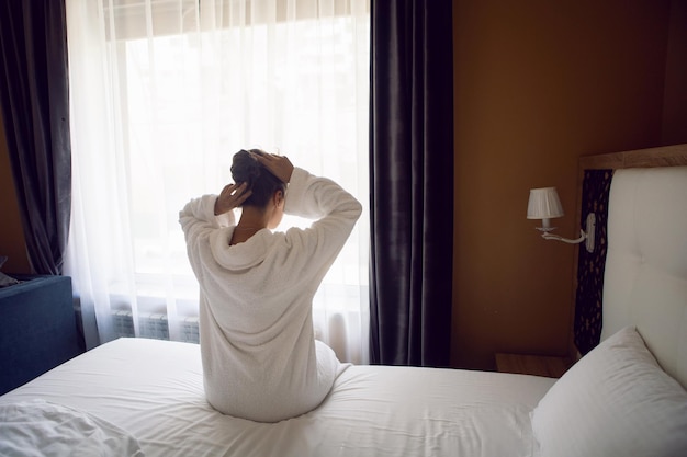 Mujer con una túnica blanca está sentada en una cama junto a la ventana en un apartamento de hotel después de una ducha