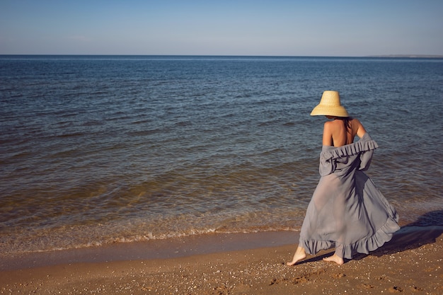 Mujer en una túnica, un traje de baño negro y un sombrero de paja se encuentra en la playa junto al mar en verano de vacaciones