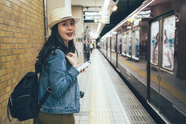 mujer turista asiática que usa teléfono móvil y mensaje de texto mientras está de pie en la plataforma subterránea en osaka, japón. feliz niña sonriente con mochila alegre emocionada por llegar al metro.