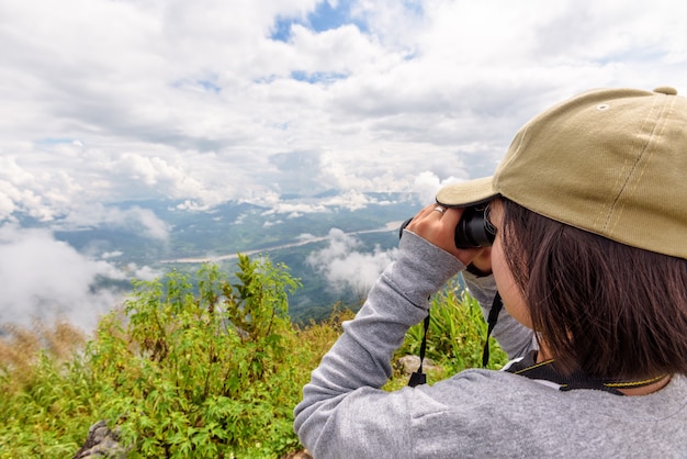 La mujer turista está usando binoculares para ver el hermoso paisaje natural del bosque del cielo y la montaña cerca del río Mekong en el mirador de Doi Pha Tang en la provincia de Chiang Rai, Tailandia