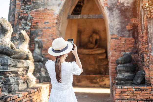 Mujer turista feliz con vestido blanco tomando fotos con un teléfono inteligente móvil, durante su visita al templo Wat Chaiwatthanaram en el Parque Histórico de Ayutthaya, concepto de viaje de verano, solo, Asia y Tailandia