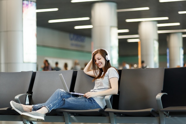 Mujer turista joven hermosa viajera con auriculares escuchando música trabajando en la computadora portátil, espera en el vestíbulo del aeropuerto internacional