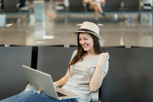 Mujer turista joven viajero alegre sentado trabajando en la computadora portátil, haciendo el gesto ganador, esperando en el vestíbulo del aeropuerto internacional