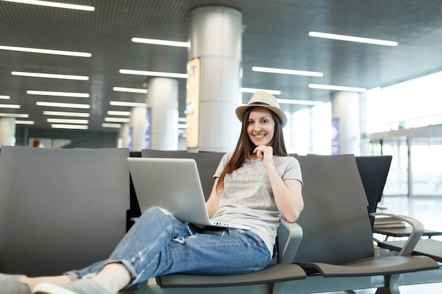 Mujer de turista joven viajero encantador con sombrero trabajando en la computadora portátil mientras espera en el vestíbulo del aeropuerto internacional