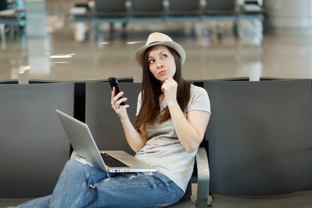 Mujer turista joven viajero pensativo con sombrero con portátil pensando, mantenga el teléfono móvil, esperando en el vestíbulo del aeropuerto internacional