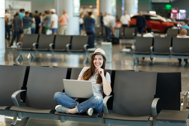 Mujer turista joven viajero pensativo con sombrero sentado con las piernas cruzadas trabajando en la computadora portátil espera en el vestíbulo del aeropuerto internacional