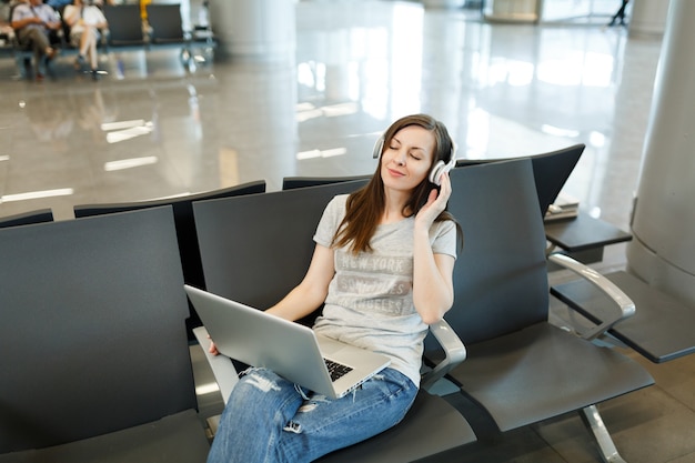 Mujer turista joven viajero relajado con auriculares escuchando música trabajando en la computadora portátil, espera en el vestíbulo del aeropuerto internacional