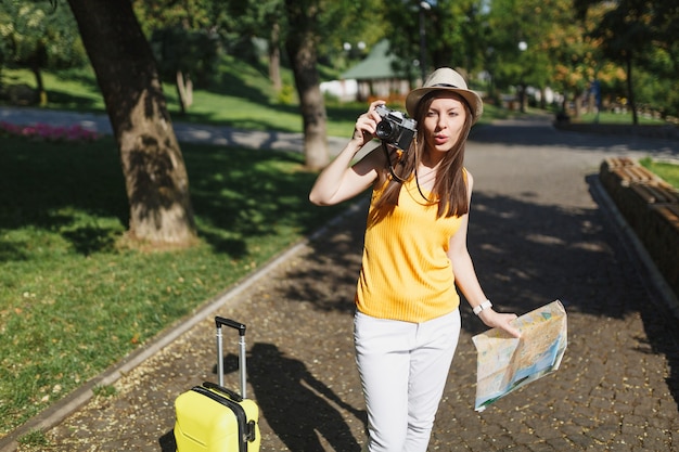 Mujer de turista joven viajero con sombrero con maleta, mapa de la ciudad tomar fotografías en la cámara de fotos retro vintage en la ciudad al aire libre. Chica viajando al extranjero para viajar en una escapada de fin de semana. Estilo de vida de viaje turístico.