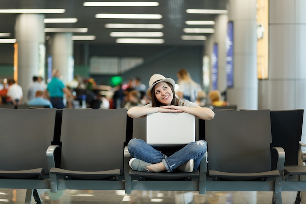 Mujer turista joven viajero soñador sentada con un portátil con las piernas cruzadas mientras espera en el vestíbulo del aeropuerto internacional