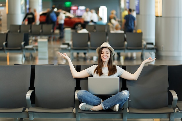 Mujer de turista joven viajero sonriente con portátil sentado con las piernas cruzadas, meditar, extender las manos, esperando en el vestíbulo del aeropuerto