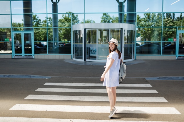 Mujer de turista joven viajero sonriente con sombrero con mochila dando la vuelta en el paso de peatones en el aeropuerto internacional