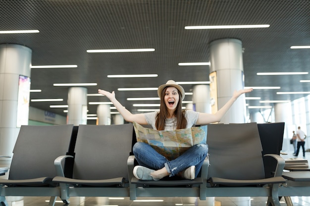 Mujer turista joven viajero sorprendido con mapa de papel sentado con las piernas cruzadas, extendiendo las manos, esperando en el vestíbulo del aeropuerto