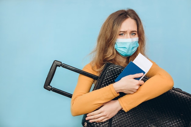 Mujer turista en una máscara médica se sienta con el equipaje en sus manos. COVID-19 brote de coronavirus. Concepto de viaje y corona. Un turista no puede irse debido a una pandemia.