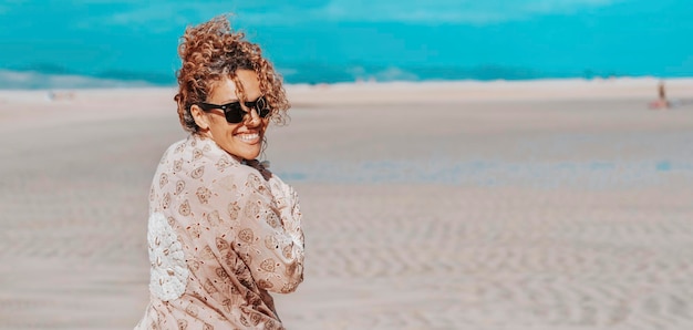 Mujer turista en la playa girando el cuerpo y mirando a la cámara sonriendo feliz Mujer adulta joven alegre disfrutar de vacaciones de verano caminando sobre la arena con el océano azul y el cielo de fondo